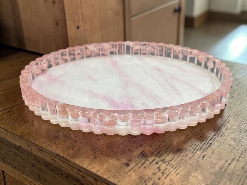 May include: A round, shallow tray with a scalloped edge, crafted from translucent resin. The base features a marbled pattern of soft pink and white swirls, while the raised, fluted rim is a clear, pale pink, creating a cohesive, layered appearance.