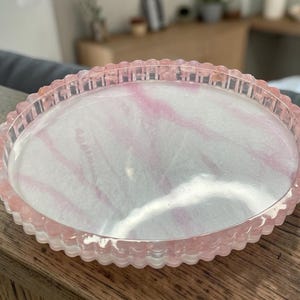 May include: A round, shallow tray with a scalloped edge, featuring a translucent pink and white marbled resin finish. The tray has a smooth, glossy surface and a raised, textured rim that follows the circular shape of the object.