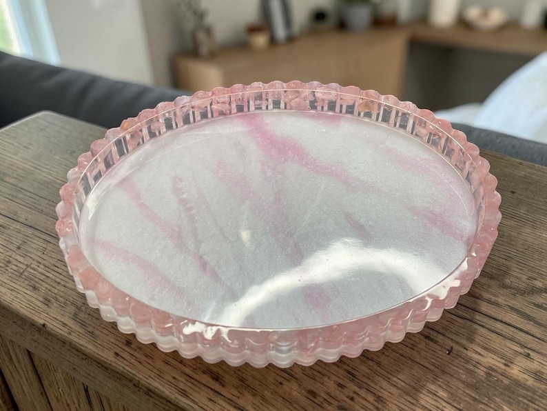 May include: A round, shallow tray with a scalloped edge, featuring a translucent pink and white marbled resin finish. The tray has a smooth, glossy surface and a raised, textured rim that follows the circular shape of the object.