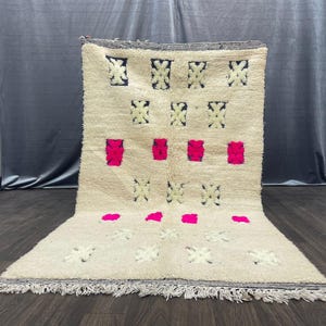 Peut inclure: Tapis rectangulaire crème avec un motif géométrique. Le tapis présente un motif répétitif de carrés noirs et crème, ainsi que des accents rectangulaires et circulaires rose vif. Le tapis a une frange sur le bord inférieur.