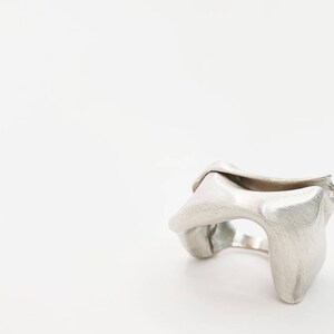 Pu&ograve; includere: Un anello color argento con un design astratto e scultoreo. L'anello ha una superficie strutturata e una forma aperta e fluida, con elementi arrotondati e curvi. L'anello sembra essere in metallo.