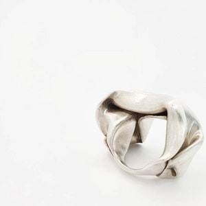 Pu&ograve; includere: Un anello d'argento con un design astratto unico. L'anello sembra essere fatto di metallo piegato, creando una forma irregolare con spazi aperti. L'anello &egrave; posto su uno sfondo bianco.