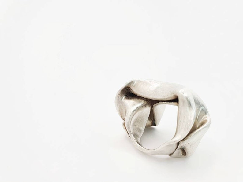 Pu&ograve; includere: Un anello d'argento con un design astratto unico. L'anello sembra essere fatto di metallo piegato, creando una forma irregolare con spazi aperti. L'anello &egrave; posto su uno sfondo bianco.