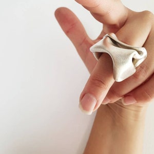 Pu&ograve; includere: Un anello color argento con un design astratto e fluido. L'anello &egrave; indossato su un dito, mettendo in risalto la sua forma e consistenza uniche. La superficie dell'anello ha una finitura opaca, che riflette delicatamente la luce. Lo sfondo &egrave; bianco.