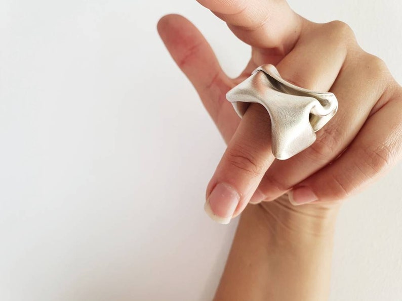 Pu&ograve; includere: Un anello color argento con un design astratto e fluido. L'anello &egrave; indossato su un dito, mettendo in risalto la sua forma e consistenza uniche. La superficie dell'anello ha una finitura opaca, che riflette delicatamente la luce. Lo sfondo &egrave; bianco.