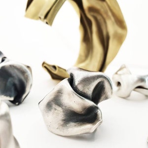 Pu&ograve; includere: Un assortimento di anelli d'argento astratti e un bracciale a polsino dorato sono esposti su uno sfondo bianco. Gli anelli hanno una forma unica e organica, con un aspetto strutturato e piegato. Il bracciale a polsino ha un design liscio e fluido.