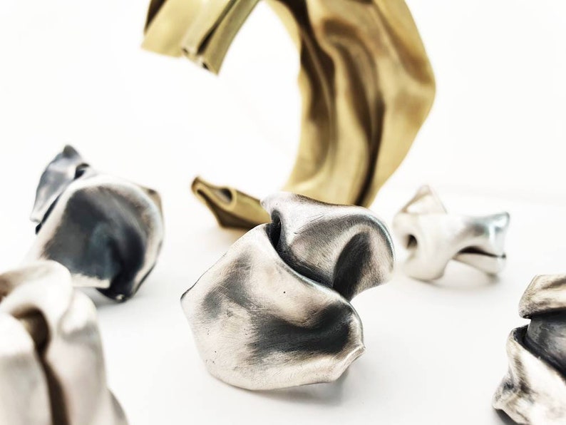 Pu&ograve; includere: Un assortimento di anelli d'argento astratti e un bracciale a polsino dorato sono esposti su uno sfondo bianco. Gli anelli hanno una forma unica e organica, con un aspetto strutturato e piegato. Il bracciale a polsino ha un design liscio e fluido.