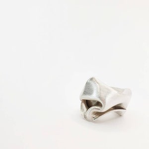 Pu&ograve; includere: Un anello d'argento con un design piegato unico. L'anello ha una superficie strutturata e una forma astratta, che sembra scolpita o modellata. L'anello &egrave; posto su uno sfondo bianco.