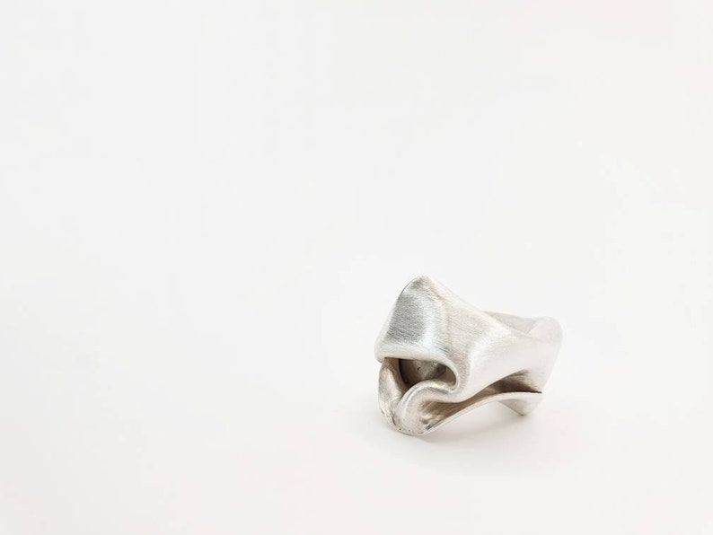 Pu&ograve; includere: Un anello d'argento con un design piegato unico. L'anello ha una superficie strutturata e una forma astratta, che sembra scolpita o modellata. L'anello &egrave; posto su uno sfondo bianco.