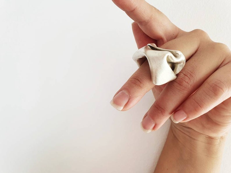 Pu&ograve; includere: Un anello color argento con un design unico e fluente. L'anello sembra essere in metallo e ha una superficie strutturata e ondulata. L'anello &egrave; indossato su un dito, mostrando la sua forma scultorea. Lo sfondo &egrave; bianco.