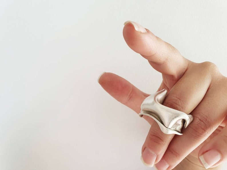 Pu&ograve; includere: Un anello color argento con un design unico e fluido. L'anello ha una forma scultorea e organica, con curve e pieghe che creano un effetto visivo dinamico. L'anello &egrave; indossato su un dito.