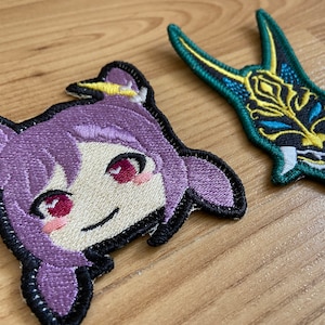 Puede incluir: Dos parches bordados. Uno presenta un personaje de estilo anime morado con una cara sonriente, y el otro es un diseño de máscara verde y dorada. Ambos parches tienen bordes negros y están sobre una superficie de madera.