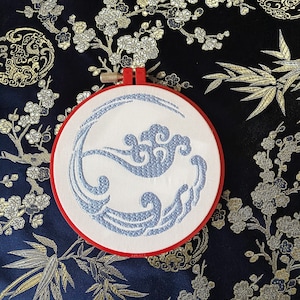 Può includere: Un ricamo blu e bianco con onde stilizzate su un tessuto bianco in un cerchio da ricamo in legno rosso.