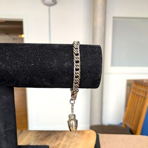 Peut inclure: Bracelet en chaîne argentée avec un petit charme décoratif. Le bracelet présente un design de chaîne gourmette et une fermeture à fermoir mousqueton. Le charme est en forme d'urne détaillée. Le bracelet est présenté sur une surface en velours noir.