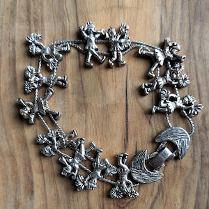 May include: Silver-toned bracelet featuring a whimsical design of figures holding onto ropes, arranged in a circular pattern. The bracelet has a clasp closure and is set against a wooden background.