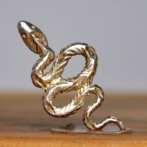Peut inclure: Figurine de serpent dorée détaillée. Le serpent est enroulé avec un corps texturé en forme de corde et une tête relevée. La figurine est montée sur une base circulaire transparente et repose sur une surface en bois.