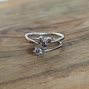 May include: A silver ring with a purple crystal Mickey Mouse head and a purple crystal star. The ring is designed to be worn on the finger.