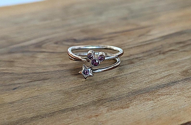 May include: A silver ring with a purple crystal Mickey Mouse head and a purple crystal star. The ring is designed to be worn on the finger.