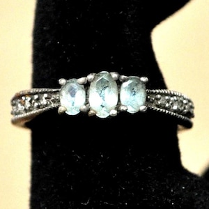 May include: A silver ring featuring three oval, light blue gemstones set in a row. The band is detailed with small, clear stones and a textured design. The ring is displayed against a black background.