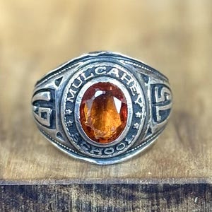 May include: A silver-toned ring with an oval, amber-colored stone at the center. The ring is engraved with the words "MULCAHEY SCHOOL" around the stone. The ring has a vintage or antique appearance.