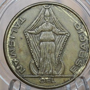 May include: A bronze-colored coin encased in a clear plastic holder. The coin features a central figure with outstretched arms, surrounded by the words "TALISMAN" and "ESPACIO". The word "DEL" is at the bottom. The coin has a vintage aesthetic.