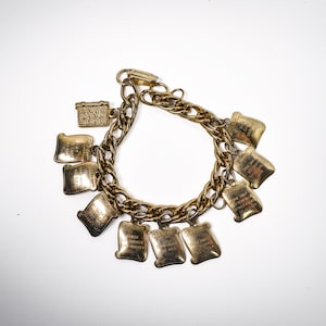 May include: A gold-tone metal charm bracelet featuring a thick curb chain link design. The bracelet is adorned with multiple rectangular charms, each engraved with text, and one small rectangular box-shaped charm attached to the main chain.