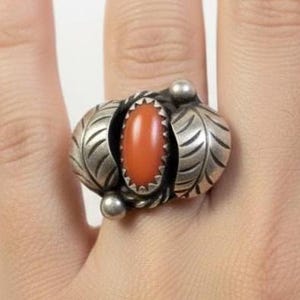 May include: A silver ring with an oval, orange-red stone at the center. The ring has a leaf design on either side of the stone and small silver balls. The ring is on a person's finger.