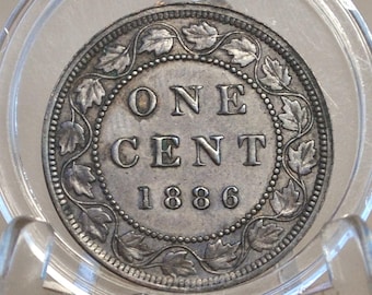 May include: A silver Canadian one-cent coin from 1886, encased in a clear protective holder. The coin features the words "ONE CENT" and the year "1886" in the center, surrounded by a detailed border of leaves and dots.