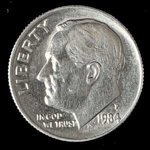 May include: A silver US dime coin with the inscription "LIBERTY" on the left side and "IN GOD WE TRUST" and "1984" on the bottom.