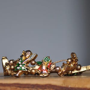 May include: A gold-toned Christmas bracelet featuring a festive design. The bracelet includes a Christmas tree, bells, and reindeer, all adorned with colorful enamel and rhinestones. A charming holiday accessory.