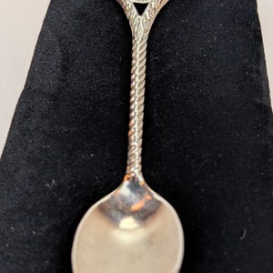 Könnte beinhalten: Ein silberner Souvenirlöffel mit gedrehtem Griff und detailliertem Emblem. Das Emblem zeigt ein goldfarbenes Bild einer Pferdekutsche in einem blau-goldenen kreisförmigen Rahmen mit den Worten "Intercourse Pennsylvania" am Rand.