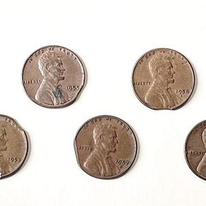 May include: Several copper-coloured Lincoln pennies, some with damaged edges, are displayed against a white backdrop. The coins show Abraham Lincoln's profile and the dates 1953, 1954, 1955, 1956, and 1958.