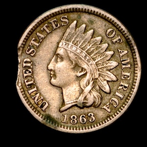 May include: An Indian Head penny from 1863. The coin features a profile of a Native American in a feathered headdress, surrounded by the words "UNITED STATES OF AMERICA" and the date. The coin is a light brown color and shows signs of wear.