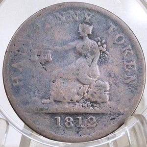 Op de afbeelding: Een antieke koperen penny token uit 1812, met een zittende figuur met weegschaal en een bundel tarwe. De munt wordt getoond in een doorzichtige houder, die de verouderde patina en ingewikkelde details laat zien. De tekst "PENNY TOKEN" is zichtbaar.