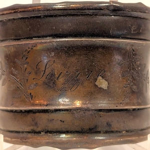 May include: A cylindrical, antique metal container with a dark, aged finish. The word "Souvenir" is etched on the side, accompanied by floral designs. The container has a ribbed texture on the top and bottom sections.