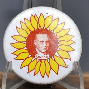 May include: A vintage white button with a yellow sunflower design surrounding a red circle. The red circle features a portrait of a man with glasses and the name "LANDON" below.