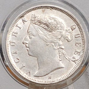 May include: A silver coin depicting Queen Victoria in profile, with the words "VICTORIA QUEEN" inscribed around the rim. The coin is housed in a clear, circular case, suggesting it's a collector's item or a piece of history.