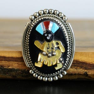 Puede incluir: Anillo ovalado plateado con una incrustación negra que representa una figura nativa americana. La figura tiene detalles turquesa, rojos y blancos, y una falda de oro amarillo. El anillo está enmarcado por un diseño de cuerda trenzada y cuentas plateadas.