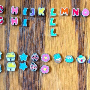 May include: Colorful alphabet letters and decorative charms arranged on a wooden surface. The letters are in various colors, and the charms feature designs like flowers, hearts, and stars. The wooden surface has vertical wood grain.