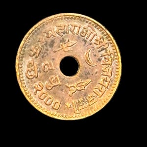 Op de afbeelding: Een bronzen munt met een centraal gat, met ingewikkelde inscripties en symbolen rond de rand. Het oppervlak van de munt vertoont tekenen van slijtage, met een datum en verschillende symbolen, waaronder een maan en een vogel.