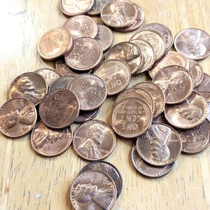 Op de afbeelding: Een verzameling koperkleurige pennies, sommige met het profiel van Abraham Lincoln, ligt verspreid op een licht houten oppervlak. De munten zijn van verschillende leeftijden, met op verschillende "One Cent" te lezen.