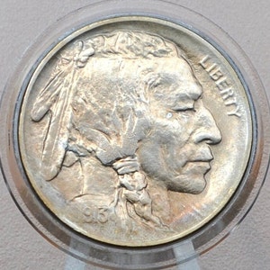 May include: A silver US Buffalo nickel coin with the year 1913 inscribed on the bottom. The coin features a profile of a Native American man with a headdress and the word "LIBERTY" inscribed on the right side.