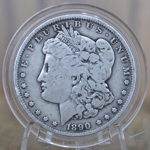 May include: A silver Morgan dollar coin with the year 1890 inscribed on the bottom. The coin features a profile of a woman with flowing hair and the inscription "E PLURIBUS UNUM" around the edge.