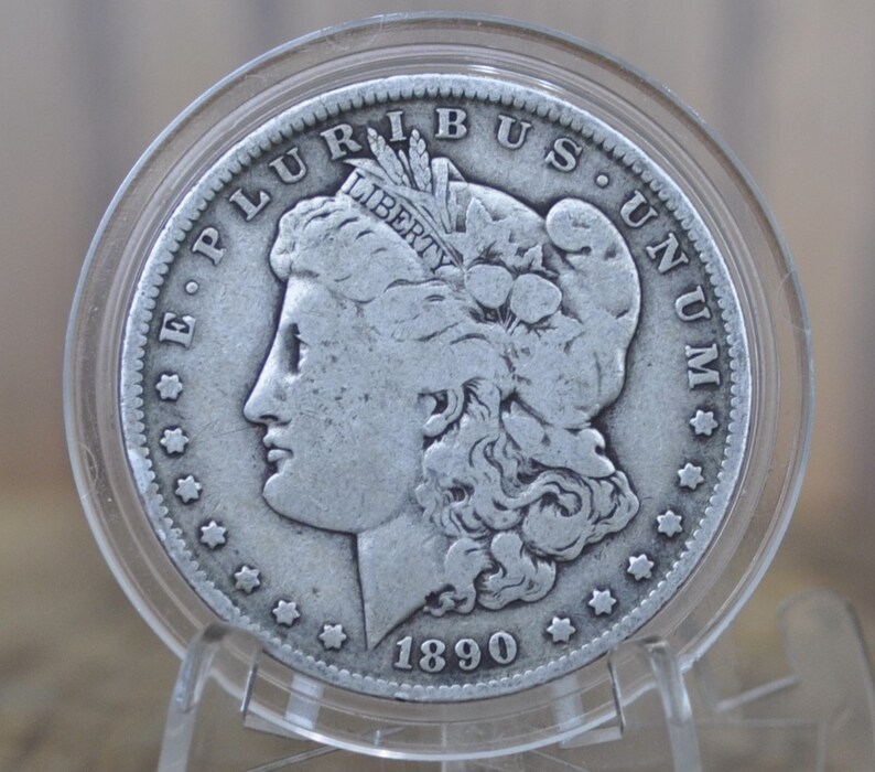 May include: A silver Morgan dollar coin with the year 1890 inscribed on the bottom. The coin features a profile of a woman with flowing hair and the inscription "E PLURIBUS UNUM" around the edge.