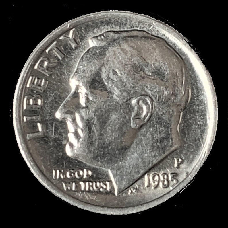 May include: A silver US dime coin with the words "Liberty" and "In God We Trust" and the year "1985" inscribed on it. The coin features a profile of President Franklin D. Roosevelt.