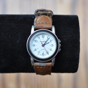 May include: A silver and brown wristwatch with a white face and black numbers. The watch face says "Aqualitic" and "Quartz".