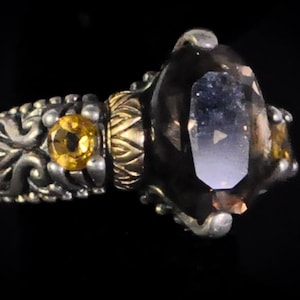 Könnte beinhalten: Ein verzierter Ring mit einem großen, facettierten, rauchquarzfarbenen Edelstein mit blauem Farbton. Der Ring hat ein silbernes Band mit goldenen Akzenten und einen kleineren gelben Edelstein. Der Ring ist vor schwarzem Hintergrund platziert.