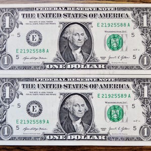 May include: Two United States one-dollar bills featuring a portrait of George Washington in the center. The bills display the text Federal Reserve Note, The United States of America, One Dollar, and serial numbers E 21925588 A and E 21925589 A.