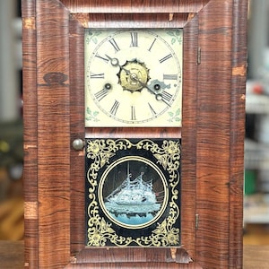 May include: A rectangular wooden clock with a dark brown grain finish. The upper section features a white clock face with black Roman numerals and ornate hands. The lower section displays a black panel with gold filigree and a ship illustration.