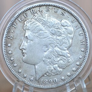 May include: A silver US Morgan dollar coin with the year 1890 and the inscription "E PLURIBUS UNUM" around the edge. The coin features a profile of Lady Liberty with flowing hair and a crown of laurel leaves.
